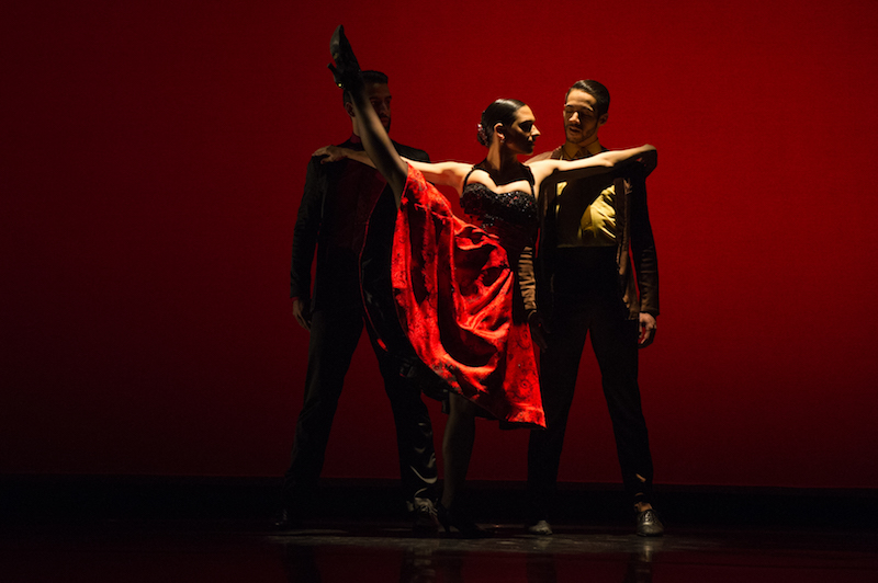 A women in a 1950s red dress extends her leg above her head while two male dancers stand behind her in the shadows